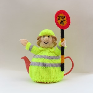 May include: A knitted teapot cosy in the shape of a person wearing a yellow and grey striped vest and a yellow hard hat. The person is holding a red stop sign with the word "STOP" written on it.