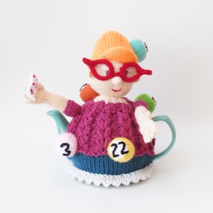 Puede incluir: Una funda para tetera de punto con forma de mujer que lleva gafas y un sombrero. La funda está decorada con bolas de colores y los números 3 y 22.