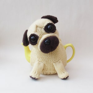 Könnte beinhalten: Ein gestrickter beigefarbener Mops-Teekannenwärmer mit braunen Ohren und einer schwarzen Nase. Der Teekannenwärmer sitzt auf einer gelben Teekanne.