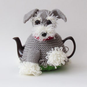 Könnte beinhalten: Ein grauer gestrickter Teekannenwärmer in Form eines Miniatur-Schnauzers. Der Wärmer hat einen weißen Pelzbesatz und eine rote Nase.