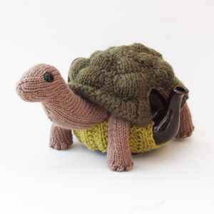 May include: A knitted green and brown turtle teapot cosy. The cosy is made of soft yarn and has a detailed shell design.