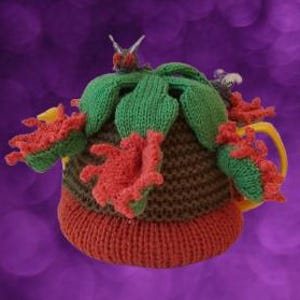 Puede incluir: Una funda de punto para tetera en color marrón y rojo, decorada con hojas verdes y flores rojas. La pieza presenta una textura de lana con forma redondeada, diseñada para cubrir una tetera, con un asa amarilla visible en el lado derecho.