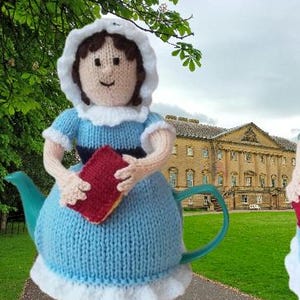 Puede incluir: Dos cubreteieras de punto con forma de mujeres con vestidos azules y gorros blancos, sosteniendo libros rojos. Las cubreteieras tienen asas turquesas y ribetes blancos. El fondo presenta un edificio señorial y hierba verde.