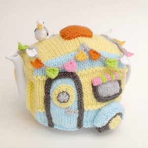 May include: A knitted teapot cosy shaped like a blue and yellow camper van with a white seagull on top. The camper has a door, windows, and a small awning with colourful bunting.