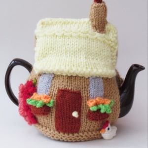 Puede incluir: Una funda de tetera de punto con forma de casa con techo amarillo, paredes de ladrillo marrón, una puerta roja y jardineras verdes. Hay un pequeño pollo de punto en el lateral.