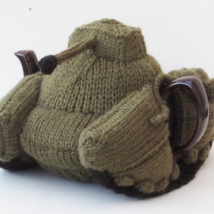May include: A green knitted teapot cosy shaped like a tank. The cosy has a brown handle and a black barrel.