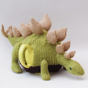 Può includere: Una teiera a forma di stegosauro lavorata a maglia nei toni del verde, marrone e beige. La teiera ha un manico giallo e una fascia marrone al centro. Il dinosauro ha placche beige sulla schiena e un piccolo occhio blu.