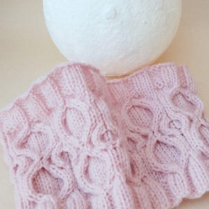 May include: A pair of light pink knitted wrist warmers with a cable knit design. The warmers are made of soft yarn and feature a repeating pattern of oval shapes. A white, textured sphere is in the background.