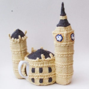 May include: A knitted teapot cozy shaped like Big Ben, the famous clock tower in London. The cozy is beige with gray details and a white porcelain teapot spout.