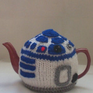 May include: A white knitted teapot cosy with blue and grey details, resembling the character R2-D2 from Star Wars. The cosy is designed to fit a standard-sized teapot.