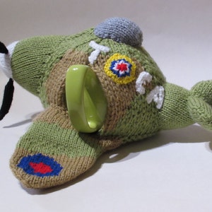 May include: A knitted green and brown airplane tea cozy with a green ceramic mug inside. The cozy has a red, white, and blue round patch on the wing and a yellow and blue round patch on the fuselage. The cozy has the word "RAF" written on it.