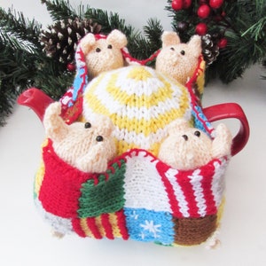 Könnte beinhalten: Eine gestrickte Teekannenhülle mit Patchwork-Muster in Rot, Grün, Weiß, Blau, Gelb und Braun. Vier kleine gestrickte Schweinefiguren schauen aus der Hülle.