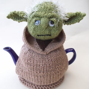 May include: A knitted Yoda tea cozy in brown with a green Yoda head on top. The Yoda head has white hair and large eyes.