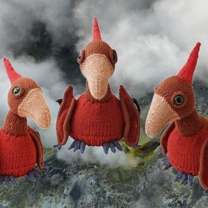 Può includere: Tre giocattoli di pterodattilo lavorati a maglia. Ognuno ha un corpo rosso, testa marrone e becco beige. Le ali sono scure e i piedi blu. Lo sfondo è un paesaggio vulcanico nuvoloso.