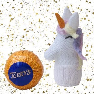 May include: A knitted white unicorn with a colorful mane and horn, next to a wrapped orange chocolate ball with a blue label that reads "Terry's". The background is white with gold star accents.