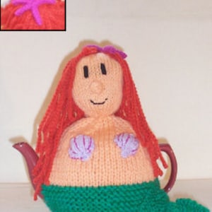 以下が含まれることがあります： 人魚の形をしたニットのティーポットカバー。カバーはピーチ色で、緑、紫、ピンクのディテールが施されています。人魚は長い赤い髪、頭に紫のヒトデ、緑の尻尾をしています。