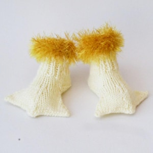 Puede incluir: Un par de botines de bebé de punto blanco con ribete peludo amarillo. Los botines tienen forma de patas de pato.