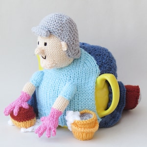 May include: A knitted tea cozy in the shape of a person wearing a blue sweater, gray hat, and pink gloves. The cozy is holding a yellow knitted mug and a brown knitted mug.