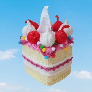 May include: A whimsical, knitted tissue box cover shaped like a cake. The cover is yellow with pink and white accents, topped with red and white knitted ornaments and colorful pom-poms. A white tissue extends from the top against a blue sky background.