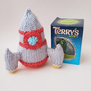 May include: A handmade, knitted rocket in light blue, red, and yellow. The rocket has a round body with red accents and a blue flower-shaped detail. A box of Terry's Chocolate Mint is next to the rocket.