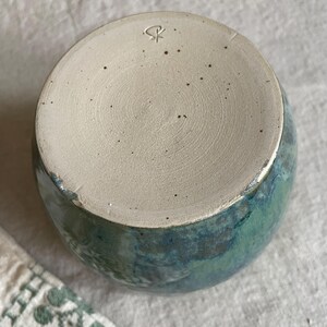 May include: A ceramic cup with a speckled beige base featuring a stamped maker's mark. The exterior displays a reactive glaze in shades of deep blue and forest green, transitioning into a smooth, unglazed circular bottom with visible clay texture.