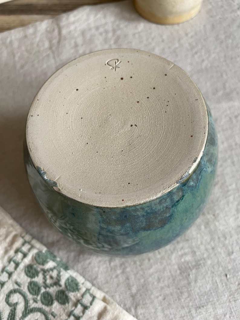 May include: A ceramic cup with a speckled beige base featuring a stamped maker's mark. The exterior displays a reactive glaze in shades of deep blue and forest green, transitioning into a smooth, unglazed circular bottom with visible clay texture.