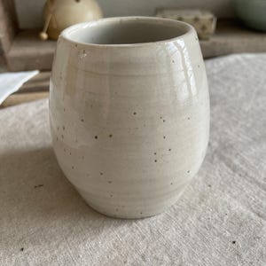 May include: A rounded ceramic tumbler with a glossy off-white glaze and visible dark brown speckles throughout the surface. The vessel features a slightly tapered base and a smooth, open rim, showcasing a handcrafted appearance with subtle horizontal ridges.