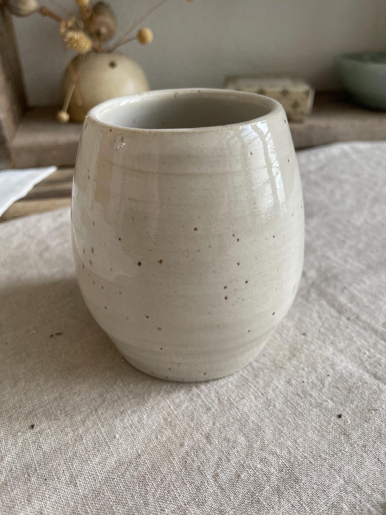 May include: A rounded ceramic tumbler with a glossy off-white glaze and visible dark brown speckles throughout the surface. The vessel features a slightly tapered base and a smooth, open rim, showcasing a handcrafted appearance with subtle horizontal ridges.