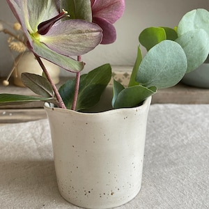 May include: A small, off-white ceramic vase with a speckled finish and an irregular, wavy rim. The vase contains a stem with dark purple hellebore flowers and a separate stem with rounded, light green eucalyptus leaves arranged inside.