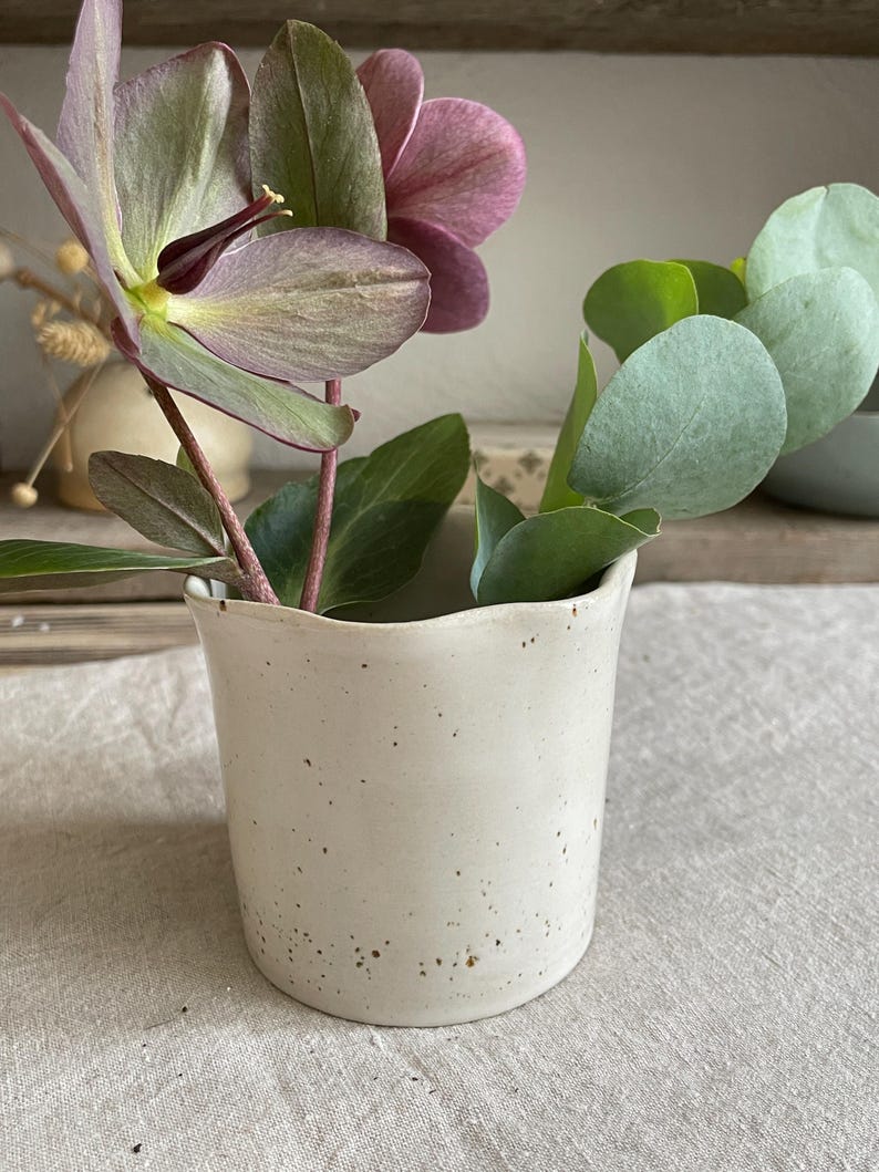 May include: A small, off-white ceramic vase with a speckled finish and an irregular, wavy rim. The vase contains a stem with dark purple hellebore flowers and a separate stem with rounded, light green eucalyptus leaves arranged inside.