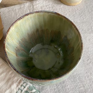 May include: A ceramic bowl with a glossy glaze featuring a blend of sage green, olive, and dark brown tones. The interior displays a radiating pattern of colors that transition from a lighter rim to a deeper, darker base with a smooth finish.