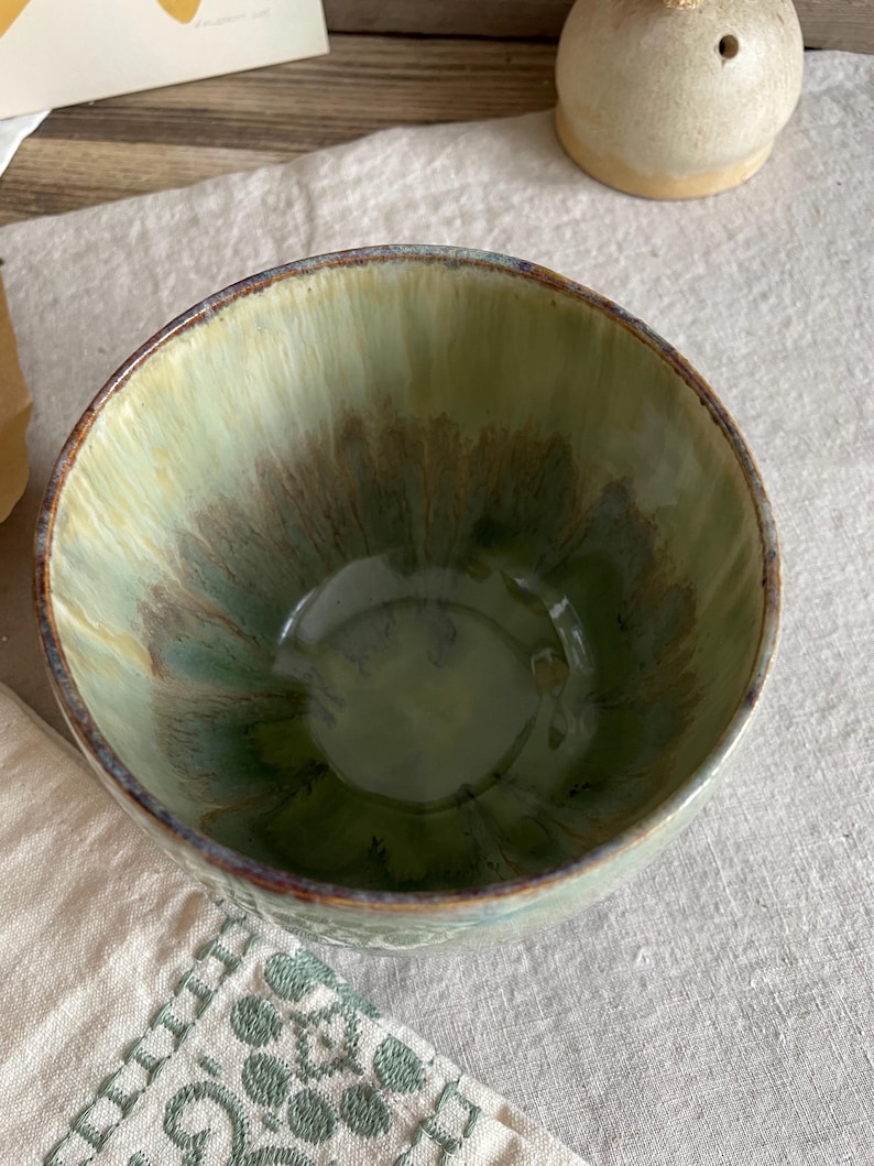 May include: A ceramic bowl with a glossy glaze featuring a blend of sage green, olive, and dark brown tones. The interior displays a radiating pattern of colors that transition from a lighter rim to a deeper, darker base with a smooth finish.