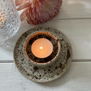 May include: A ceramic candle holder with a lit tea light candle. The holder is speckled with brown spots and sits on a matching plate. A pink flower and a textured glass candle holder are in the background.