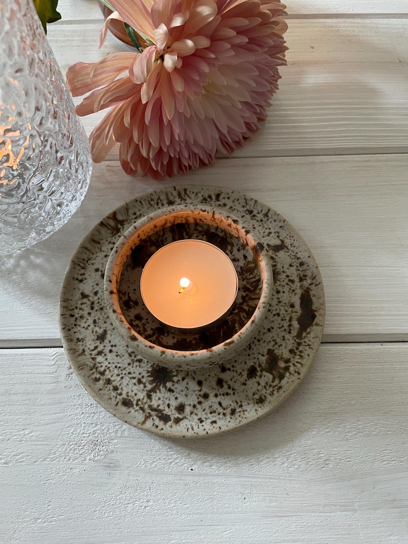 May include: A ceramic candle holder with a lit tea light candle. The holder is speckled with brown spots and sits on a matching plate. A pink flower and a textured glass candle holder are in the background.