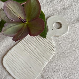 Könnte beinhalten: Ein weißes Servierbrett aus Keramik mit einer strukturierten Oberfläche, die feine Wellenlinien aufweist, und einem runden Griffausschnitt. Eine einzelne Nieswurzblüte mit violetten und grünen Blütenblättern und dunkelgrünen Blättern liegt darauf.
