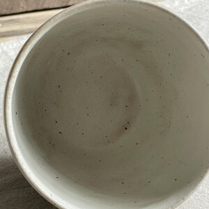 May include: A ceramic cup with a speckled off-white glaze. The interior of the cup features a smooth, light-colored surface with small dark brown flecks distributed throughout the material, showing a simple, open, and circular rim design.