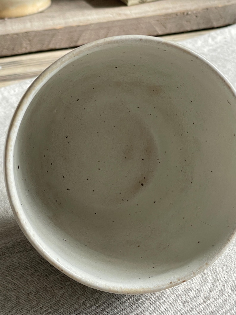 May include: A ceramic cup with a speckled off-white glaze. The interior of the cup features a smooth, light-colored surface with small dark brown flecks distributed throughout the material, showing a simple, open, and circular rim design.