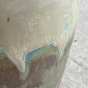 May include: A ceramic vessel featuring a reactive glaze with a smooth, creamy beige upper section that transitions into a textured, mottled base of deep brown, sage green, and teal blue, with small dark brown speckles visible throughout the surface.