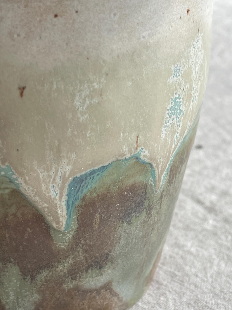 May include: A ceramic vessel featuring a reactive glaze with a smooth, creamy beige upper section that transitions into a textured, mottled base of deep brown, sage green, and teal blue, with small dark brown speckles visible throughout the surface.