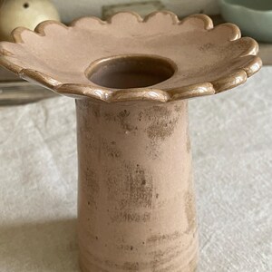 May include: A dusty pink ceramic vase with a wide, scalloped, flower-shaped rim and a cylindrical body. The surface features a textured, mottled finish with darker brown patches, giving it a weathered appearance. The center has a circular opening.