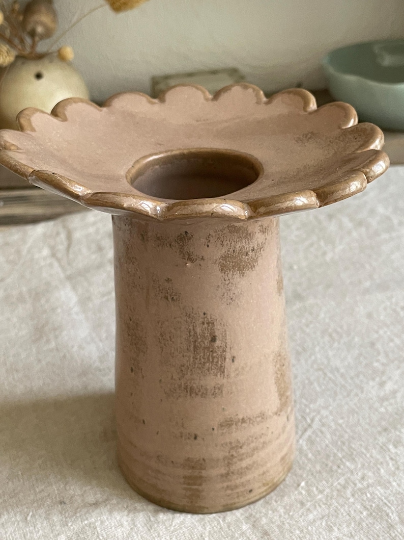 May include: A dusty pink ceramic vase with a wide, scalloped, flower-shaped rim and a cylindrical body. The surface features a textured, mottled finish with darker brown patches, giving it a weathered appearance. The center has a circular opening.