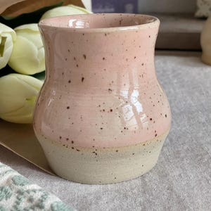 Könnte beinhalten: Eine kleine, handgedrehte Keramikvase mit ausgestelltem Rand und bauchigem Körper. Der obere Teil ist mit einer glänzenden, hellrosa Glasur mit dunkelbraunen Sprenkeln versehen, während die Basis aus unglasiertem, mattem, beigem Steinzeug besteht.