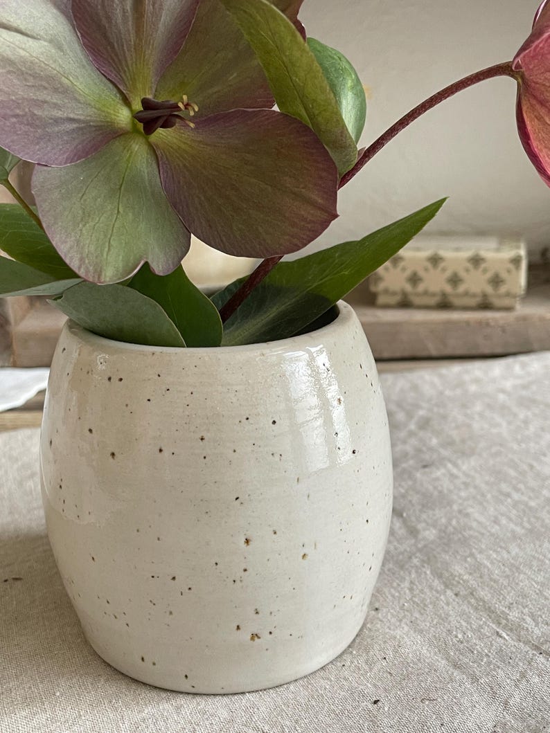 May include: A small, barrel-shaped ceramic vase with a glossy off-white glaze and dark brown speckles throughout. The vase contains several hellebore flowers with muted purple and green petals and dark green leaves extending from the opening.