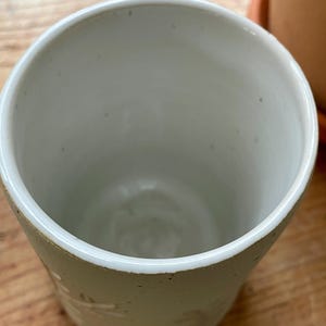 Pu&ograve; includere: Una tazza in ceramica fatta a mano, bianco sporco, con una superficie leggermente strutturata. La tazza ha un'ampia apertura e un interno liscio e bianco. Sottili motivi floreali o a forma di foglia sono visibili all'esterno.