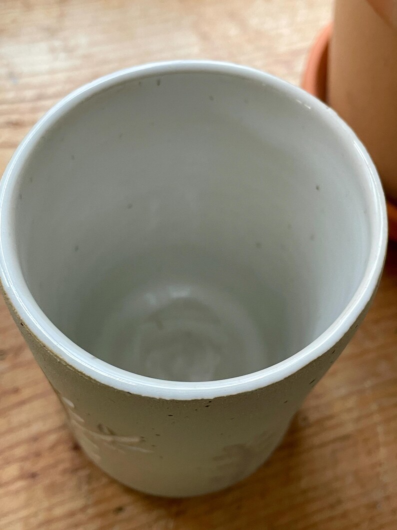 Pu&ograve; includere: Una tazza in ceramica fatta a mano, bianco sporco, con una superficie leggermente strutturata. La tazza ha un'ampia apertura e un interno liscio e bianco. Sottili motivi floreali o a forma di foglia sono visibili all'esterno.