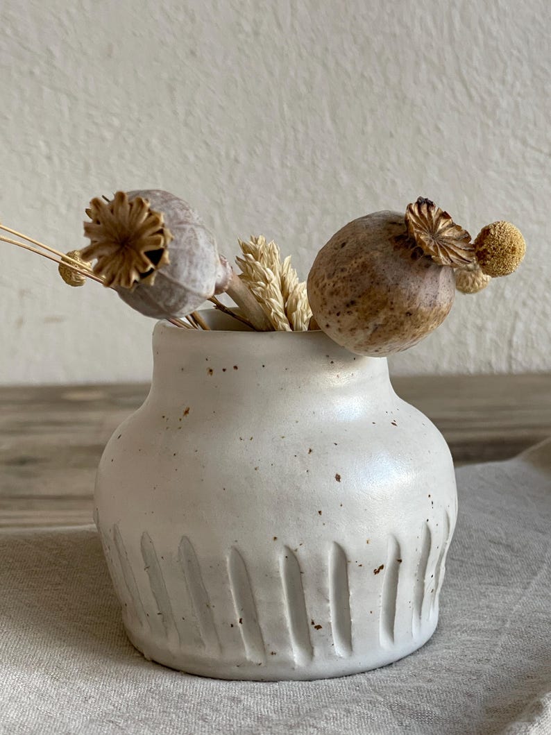 May include: A small, off-white ceramic vase with vertical grooves near the base, holding dried seed pods and wheat stalks. The vase has a speckled texture and a rounded shape, creating a rustic, minimalist aesthetic. The arrangement is set against a neutral background.