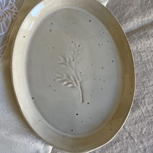 Könnte beinhalten: Eine ovale Keramikplatte mit einer glänzenden cremeweißen Glasur und gesprenkelter Oberfläche. In der Mitte der Platte befindet sich ein erhabenes, geprägtes botanisches Zweigmotiv mit zarten Blättern, das eine subtile Textur erzeugt.