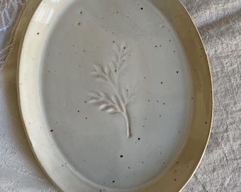 Può includere: Un piatto da portata ovale in ceramica con una smaltatura lucida color bianco sporco e una finitura maculata. Il centro del piatto presenta un motivo botanico in rilievo con foglie delicate, creando una texture sottile sulla superficie.