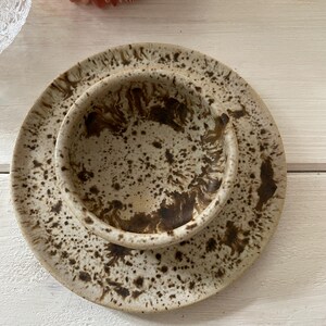 May include: A ceramic bowl and plate set with a speckled, earthy design. The bowl sits atop the plate, both featuring a beige base with brown speckles. The set is ideal for serving food and adds a rustic touch to any table setting.