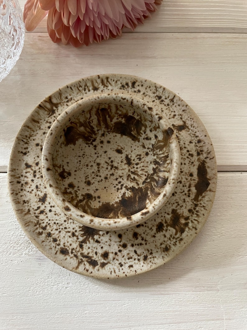 May include: A ceramic bowl and plate set with a speckled, earthy design. The bowl sits atop the plate, both featuring a beige base with brown speckles. The set is ideal for serving food and adds a rustic touch to any table setting.